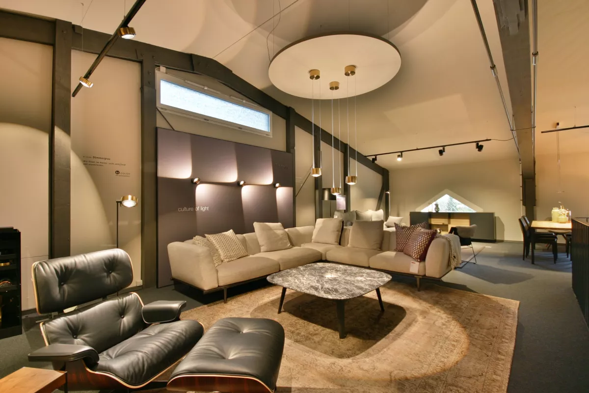 Showroom Wohnbereich mit Sofa, Sessel, Salontisch, Teppich, grosse runde Lampe an der Decke mit drei kleinen Lichtern, die herunterhängen und weitere Deckenlampen