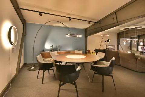 Showroom Essbereich mit runden Holzesstisch und 6 moderen Stühlen, Stehlampe beleuchtet den Tisch, rundes Licht an der Wand